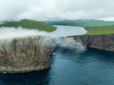 Aerial view of the ethereal lake seemingly suspended above the ocean's edge, embraced by dramatic cliffs shrouded in mist and green hills, Sorvagsvatn lake, Vagar, Faroe Islands.