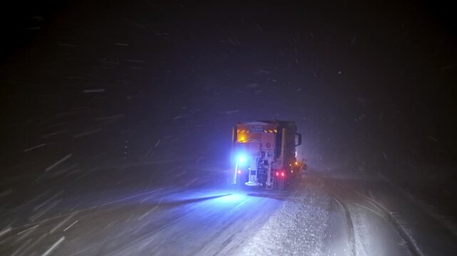 View from inside a vehicle following a snowplow clearing a snow-covered road at night. Blowing snow, flashing warning lights and low visibility show dangerous winter driving and road maintenance opera