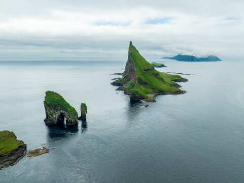 Aerial view of verdant isles rise majestically from the sea, their rugged cliffs and emerald slopes contrasting with the moody, overcast sky, Tindholmur, Faroe Islands.