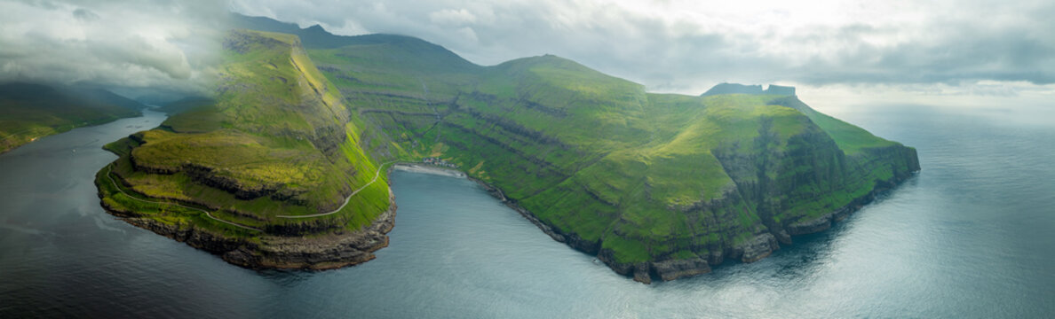 Aerial view of dramatic cliffs meet the moody sea, where a small village nestles in a cove beneath the green slopes, Tjornuvik, Streymoy, Faroe Islands.