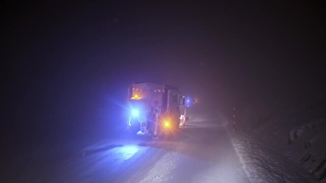 View from inside a vehicle following a snowplow clearing a snow-covered road at night. Blowing snow, flashing warning lights and low visibility show dangerous winter driving and road maintenance opera