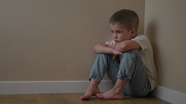 Child curled up low in room corner, hugging knees tightly. Sadness reflected in child eyes and face. Sitting posture stays frozen. Emotion and corner silence surround child with still loneliness.