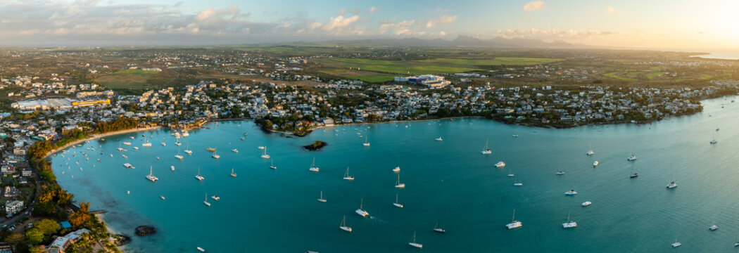 Aerial view of turquoise waters speckled with white sailboats meeting a vibrant coastline lined with buildings and greenery, Grand Baie, Riviere du Rempart District, Mauritius.