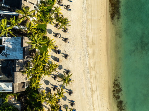 Aerial view of thatched umbrellas dotting the sandy beach bordered by turquoise waters and buildings, Shangri La, Trou d'Eau Douce, Mauritius.