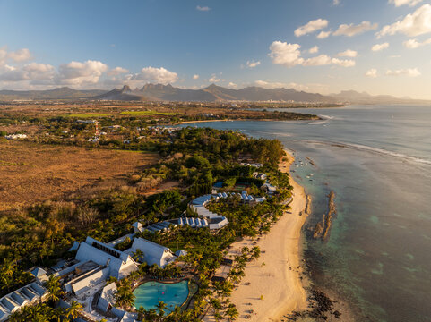 Aerial view of the coastline meeting the turquoise sea, with buildings nestled among lush greenery, set against a backdrop of distant mountains, Grand Baie, Riviere du Rempart District, Mauritius.