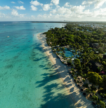 Aerial view of the coastline where the turquoise ocean meets the sandy beach, fringed by lush green trees casting long shadows, Triolet, Pamplemousses, Mauritius.
