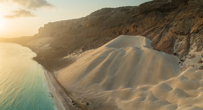 Aerial view of the golden dunes meeting the turquoise sea under a hazy sky, nestled against rugged cliffs, Arher dunes, Socotra, Yemen.