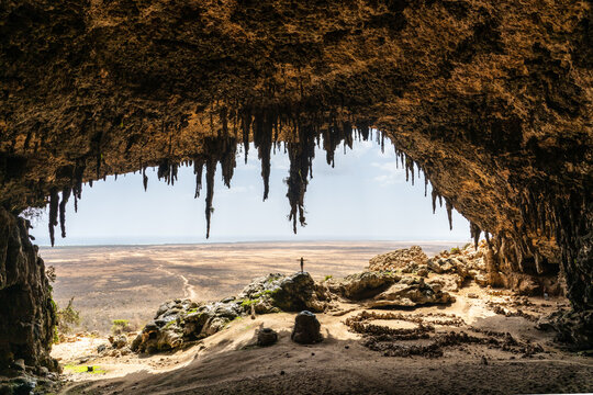 View of a person stands atop a rocky outcrop in a vast cave, its ceiling adorned with stalactites, framing a distant arid landscape, Socotra, Yemen.