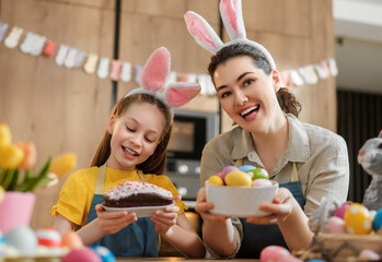 Happy Mother and Daughter Celebrating Easter in Kitchen with Bunny Ears and Colorful Eggs