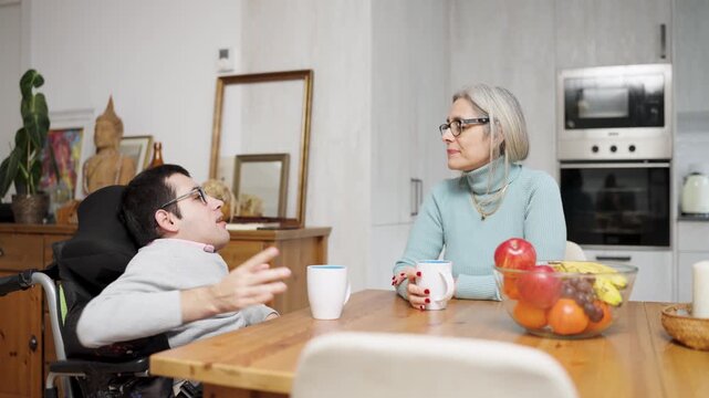 Loving son with a physical condition sitting in a wheelchair while having an engaging conversation with his senior mother at the kitchen table