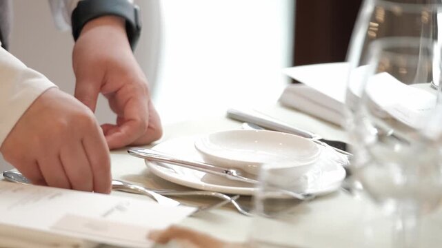 Close up of a waiter in a white shirt setting a formal dinner table with plates and cutlery. Static shot.