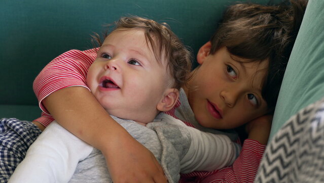 Older sibling cuddling baby on the couch, tender moment with the baby looking joyful and expressive, siblings sharing an intimate bonding experience in a cozy home setting