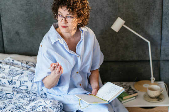 Thoughtful White woman in her 40s with glasses pausing journaling in bedroom