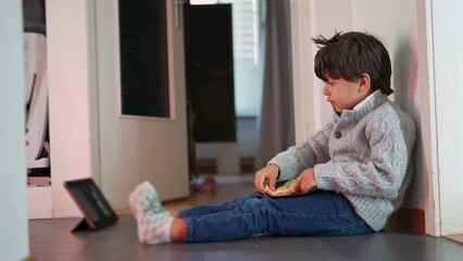 Fototapeta premium Young boy sitting on floor, eating bread, watching tablet, relaxed and engaged, casual home setting, child enjoying snack, focused expression, indoor activity