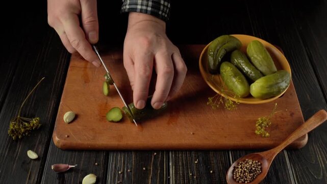 Hands slicing pickles on a wooden cutting board with a bowl of whole pickles and spices arranged nearby in a cozy kitchen setting