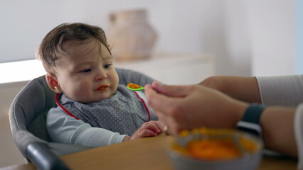 Close-up of baby trying solid food for the first time, slightly reluctant expression, spoon near mouth, home setting, gentle introduction to new flavors, nurturing care