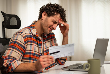 Stressed man using laptop and credit card to pay his financial bills online while sitting at home. He is having a headache and worried expression while managing his budget and expenses.	
