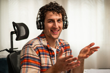 Close up photo of happy young man wearing wireless headphones, talking and gesturing while attending video call from his cozy home.