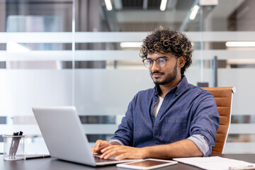Concentrated young Indian man wearing glasses sitting at a desk in the office and working on a...