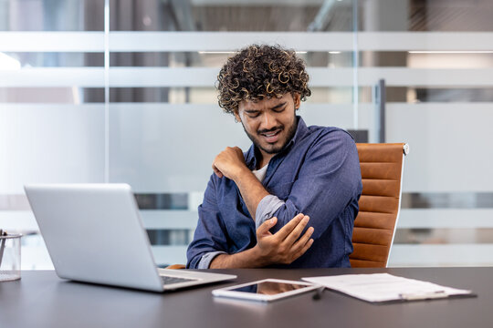 A young Indian man, twisted in pain, sits in the office and holds his elbow with his hand