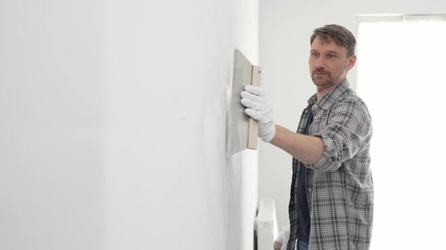 Male construction worker wearing protective gloves and plaid shirt, spreading plaster smoothly across wall surface using professional drywall taping knife during renovation project. Portrait view
