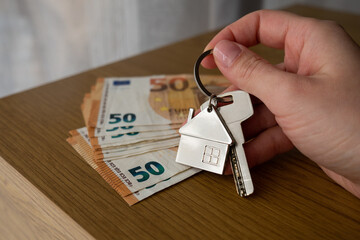 Female hand holding house keychain with euro bills on table, real estate investment and property ownership.