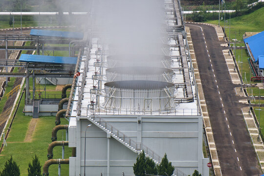 Aerial view of a geothermal power plant with cooling towers emitting steam, surrounded by green landscape and industrial infrastructure.