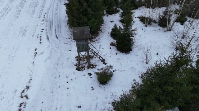 Aerial drone footage of a wooden hunting stand (high seat) surrounded by snow-covered forest in Podlaskie region, northeastern Poland. Winter landscape with evergreen trees and rural wilderness scener