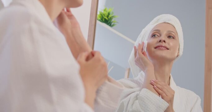 Beautiful woman looking at her face reflection in mirror, applying cream skin, wearing white bathrobe, towel wrapped, smiling happily and looking relaxed, personal skincare or beauty routine to clean 