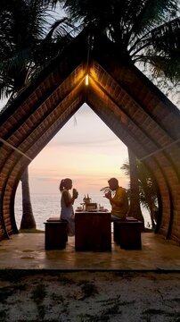 Enjoy a romantic dinner at a charming beachfront pavilon in Koh Mak Thailand. The couple savors delicious cuisine while the sun sets over the ocean, creating a magical atmosphere.