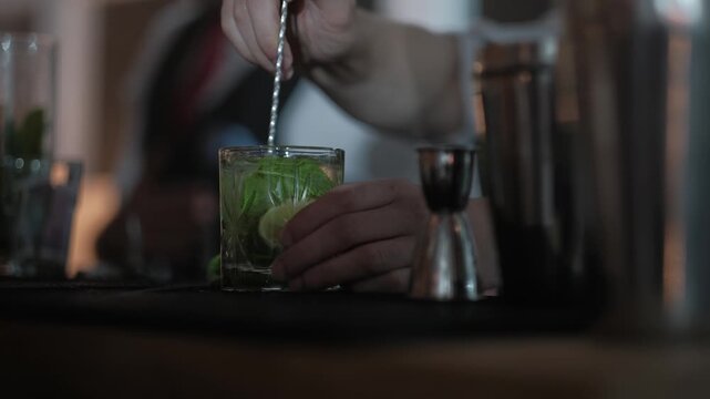 Professional bartender in a white shirt and black vest stirring a mojito cocktail with a long bar spoon