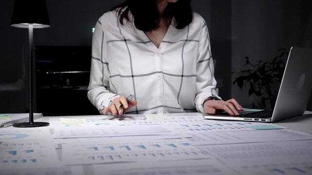 Woman working late in an office, reviewing numerous reports and typing on a laptop, demonstrating diligent attention to detail and a strong work ethic