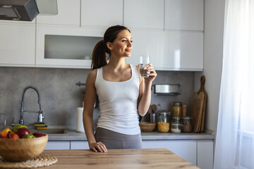 Caucasian beautiful woman in sportswear drink water after exercise. Young thirsty active sport girl takes a sips of clean mineral natural in cup after workout for health care in kitchen in house. © Jelena Stanojkovic