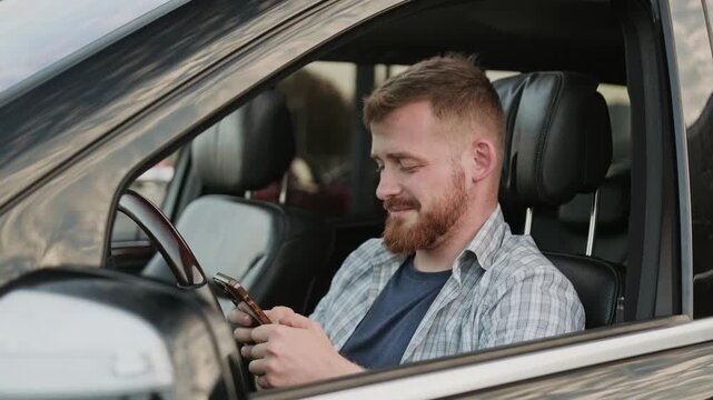 Smiling young man looking at smartphone screen, sitting in the car. Slow motion