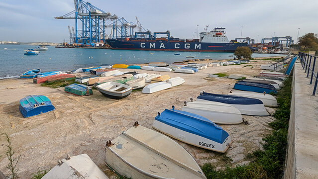 Colorful upside down dinghies on concrete shoreline beside calm bay, with container ship and blue cranes in Birzebbuga Freeport Malta: January 4, 2026 - Birzebugga, Malta