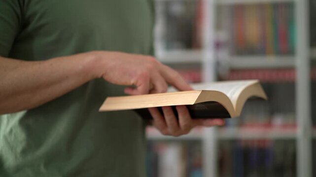 Man Holding Open Paperback Book, CloseUp Of Hands Flipping Pages With Soft Shelf Background, Textured Paper