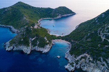 Obraz premium A serene blue hour aerial photograph of Porto Timoni's twin bays in Corfu, Greece. A single sailboat rests in the calm cove, nestled between rugged, green cliffs after sunset.