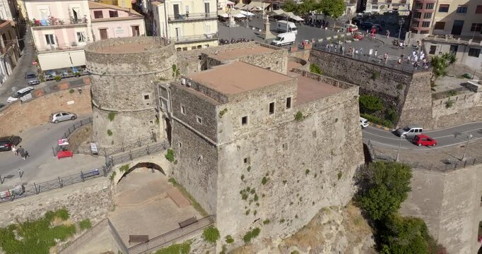 Aerial closeup of the main entrance to a medieval stone fortress. The shot captures a robust circular tower with narrow arrow slits and the gated entrance with a bridge. It's located in Pizzo, Italy.
