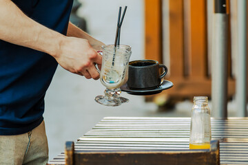 Moment of routine service - a hand lifts a glass with remnants of frothy drink, alongside a dark coffee cup and an empty bottle on the table. The sunny outdoor setting conveys a relaxed, everyday atmo
