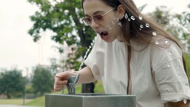 Woman fumbles stream from tricky sidewalk fountain under leaves. Casual shirt shifts in hydration fail.