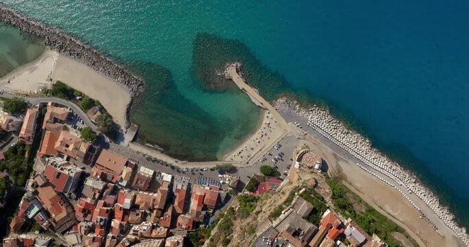 Aerial zenithal view of Pizzo Calabro beach, located on the Coast of the Gods, Calabria, Italy. A stone pier and a small beach divide the turquoise water from the dense cluster of historic buildings.