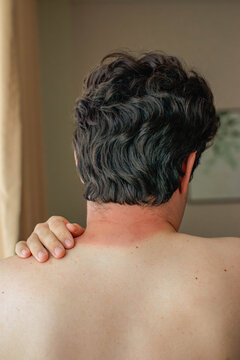 Rear view of a man with dark curly hair, sunburn on the back of his neck, left hand resting on shoulder, neutral indoor background