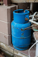 Obraz premium Old blue metal gas cylinder with rust and scratches standing outdoors near cables and wall, symbolizing fuel storage, industrial equipment and safety concept