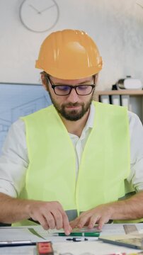 Good-looking skilled experienced bearded engineer-constructor in special workwear and protective helmet drawing line on paper project with pen and protractor in workroom