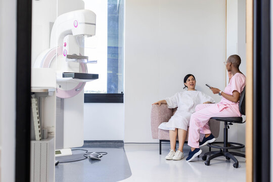 Female patient and female tech sitting in mammography room in gown using tablet, copy space