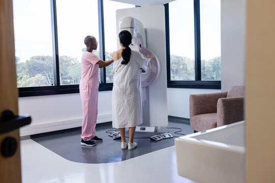African American pink-scrub tech positioning Asian woman patient in white gown at mammography unit