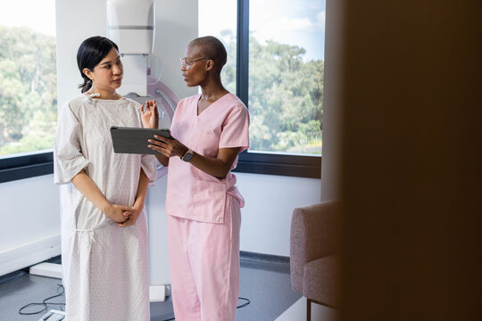 Asian patient in gown and African American nurse, women using tablet by window, pointing at scanner