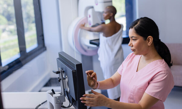 Diverse female patient in white gown, female tech in pink scrubs operating mammogram, copy space