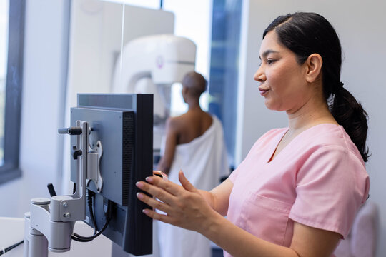 African American woman tech in pink scrubs using monitor in mammo room, patient draped, copy space