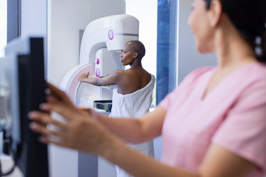 Diverse female patient in gown and tech placing breast on mammogram in clinic while using console
